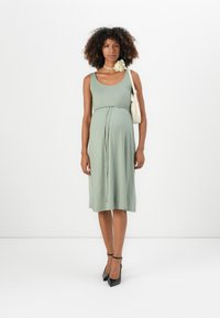 Mulher com cabelo encaracolado, vestindo um vestido sem mangas de um verde claro, sapatos de salto pretos, carregando uma bolsa branca de ombro e usando um colar com uma flor amarela.