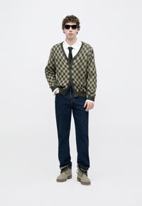 Πράσινη και μπεζ ζακέτα με καρό σχέδιο, λευκό πουκάμισο, μαύρη γραβάτα, μπλε τζιν και γκρι μπότες, με γυαλιά ηλίου. Υφασμάτινη υφή και άνετη εφαρμογή.