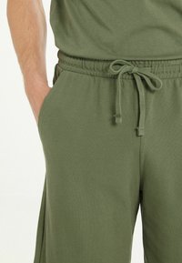 Pantalones de chándal verde oliva con cinturilla elástica, cordón ajustable, bolsillos laterales y tela texturizada. Diseño sencillo con un ajuste relajado.