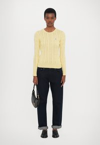 Suéter amarelo de malha com punhos canelados, calças de ganga de corte largo em azul escuro, dobradas no tornozelo, e uma pequena bolsa preta.