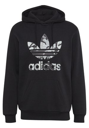 Osoba ubrana w czarną bluzę z kapturem Adidas z logo, beżowe spodnie sztruksowe podwinięte u dołów oraz białe buty sportowe Adidas z czarnymi paskami.