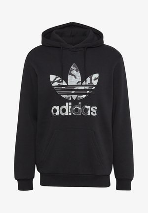 Person trägt schwarzen Adidas Hoodie mit Logo, beige Cordhosen mit aufgekrempelten Bündchen und weiße Adidas-Sneaker mit schwarzen Streifen.