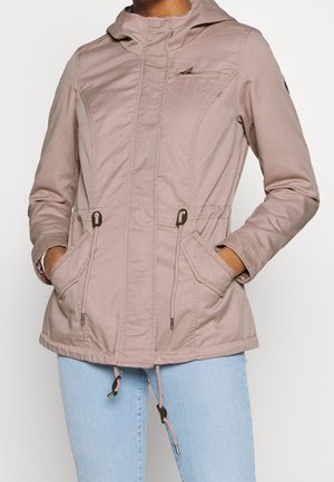 Femme portant une veste à capuche rose clair avec cordons, poche poitrine zippée et mains dans les poches avant, associée à un jean bleu clair.
