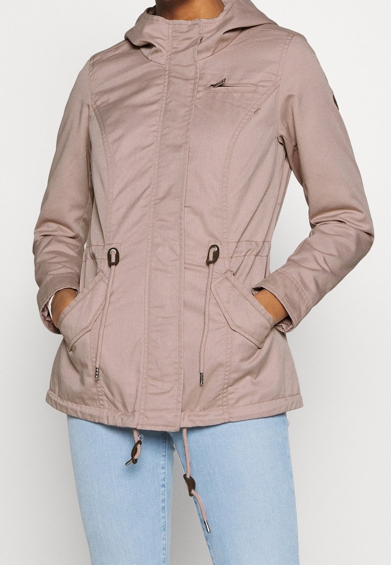 Femme portant une veste à capuche rose clair avec cordons, poche poitrine zippée et mains dans les poches avant, associée à un jean bleu clair.