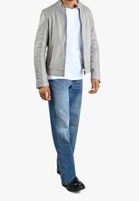 Uomo che indossa una giacca di pelle grigia chiara, una maglietta bianca, jeans blu e scarpe nere, in piedi davanti a uno sfondo bianco uniforme.