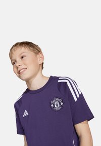 Lila Manchester United T-Shirt aus strukturiertem Stoff, mit weißen Adidas-Streifen an den Ärmeln und gesticktem Vereinslogo auf der Brust.