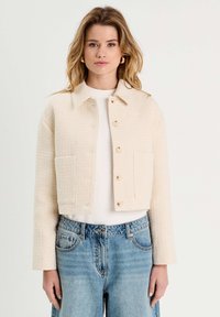 Gekürzte beige Jacke aus strukturiertem Stoff, mit Knopfverschluss, spitzem Kragen und zwei Seitentaschen, kombiniert mit hellblauen Jeans.