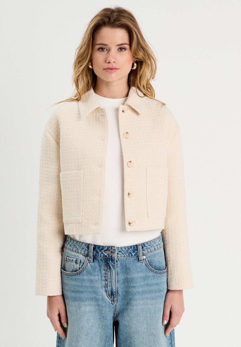 Gekürzte beige Jacke aus strukturiertem Stoff, mit Knopfverschluss, spitzem Kragen und zwei Seitentaschen, kombiniert mit hellblauen Jeans.