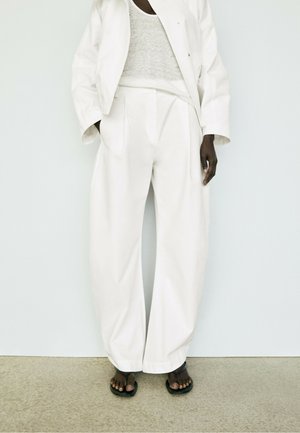 Persona che indossa pantaloni bianchi larghi, una giacca bianca, una maglia grigio chiaro e sandali neri, in piedi contro un muro semplice.