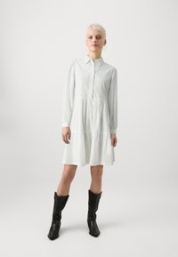 Lys grønn, langermet kjole med knappestolpe foran, lagdelt design og teksturert stoff; kombinert med svarte, dekorative cowboy-støvler.