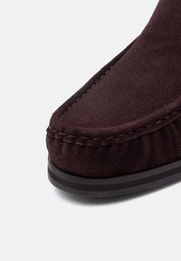 Κοντινή λήψη σκούρου καφέ μοκασίνι σουέντ, που δείχνει λεπτομερή ραφή και μαύρη σόλα σε λευκό φόντο.