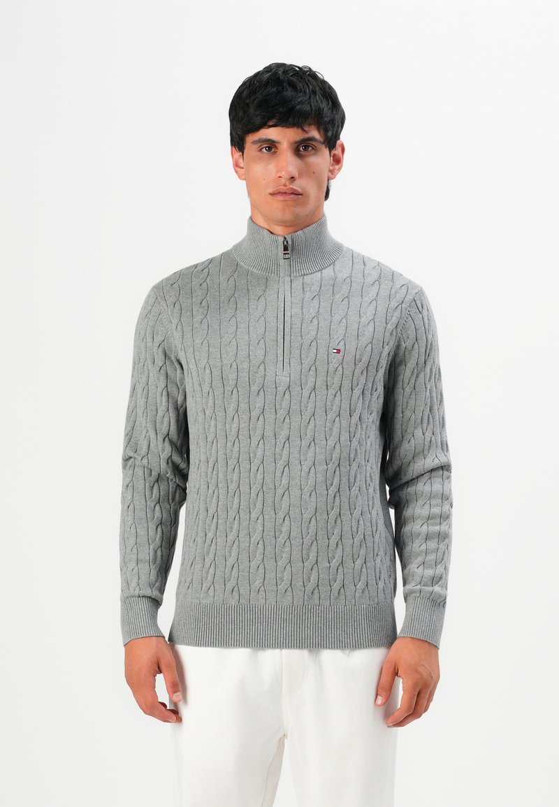 Mann in einem grauen Pullover mit Kabelstrick und Viertel-Reißverschluss, mit einem kleinen Logo auf der Brust, trägt weiße Hose und steht vor einem weißen Hintergrund.