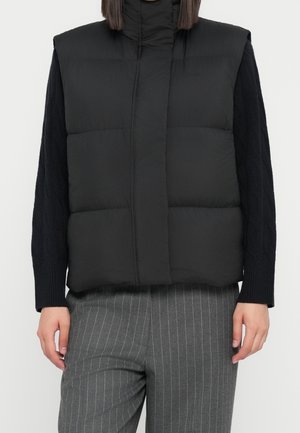 Kvinde iført en sort dunvest over en sort strikket sweater og grå bukser med nålestriber, stående foran en ensfarvet baggrund.