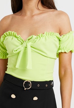 Femme portant un crop top vert citron à épaules dénudées avec volants et nœud, associé à une jupe taille haute noire avec boutons dorés et ceinture.