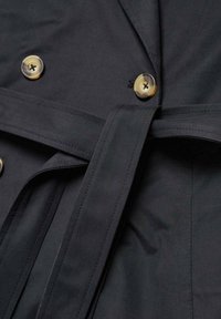 Černý trench coat z látky s zlatými knoflíky a odpovídajícím pásem. Hladká textura s prošíváním na přední straně.