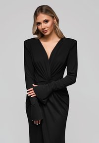 Abito lungo nero a maniche lunghe con profondo scollo a V, dettaglio in vita annodato e maniche svasate. Tessuto morbido con una consistenza liscia.