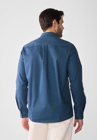 Camisa de manga larga azul marino con cuello abotonado, pliegue en la parte posterior y dobladillo redondeado, hecha de suave tela de algodón y con puños abotonados.