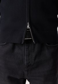 Svart zip-up genser med ribbestrikket kant og mansjetter, sammen med mørk denim jeans. Glidelåsen er metallisk og plassert i midten foran.