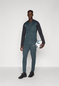 Nikeské teplákové súpravy v tmavo smaragdovej farbe s čiernymi rukávmi, obsahujúce zipsovú bundu a slim-fit nohavice. Hráč drží futbal v jednej ruke. Čierne tenisky.