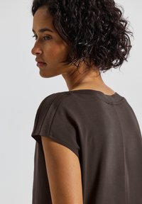 Haut marron à manches courtes avec une texture lisse, présentant un col rond et des détails subtils aux épaules, affiché de profil.