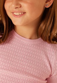 Rosa gestricktes Oberteil mit einem strukturierten Muster und geripptem Ausschnitt. Kleine Regenbogen- und Stern-Ohrringe sind am Ohr sichtbar.