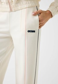 Nahaufnahme von cremefarbenen Jogginghosen mit pinken und schwarzen Seitestreifen, GUESS-Logo am Oberschenkel und einer Hand, die in der Tasche ruht.