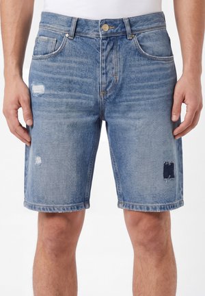 Hombre con pantalones cortos de mezclilla azul con detalles desgastados y remendados, de pie con las manos relajadas a los lados contra un fondo blanco.