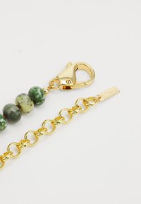 Χρυσή αλυσίδα με στρογγυλές πράσινες και γεμάτες αστιχίες χάντρες, με κούμπωμα καβουριού και μικρή ετικέτα με χαραγμένο το "élou." Λειασμένη επιφάνεια, γυαλιστερός μέταλλος.