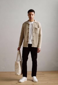 Gilet en tricot beige avec des boutons marron, t-shirt blanc, pantalon noir et baskets blanches. Sac fourre-tout en coton décontracté à la main.