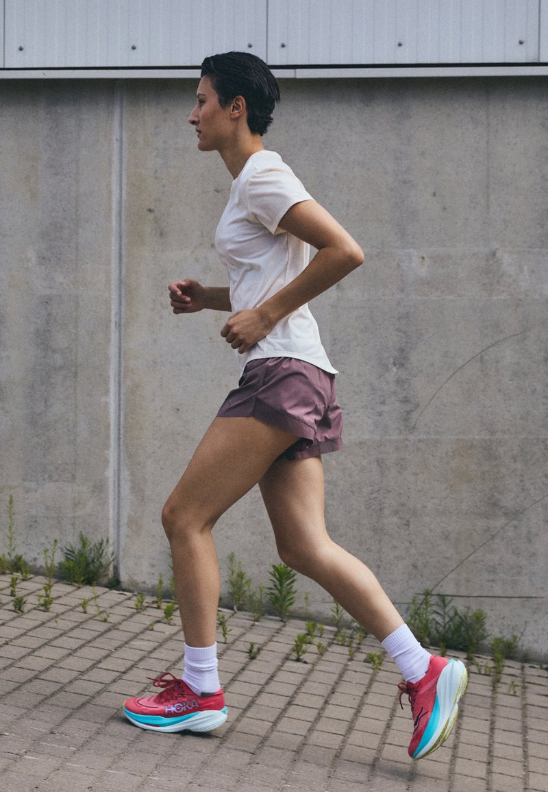 Tenue de sport comprenant un t-shirt ajusté blanc, un short violet avec un ourlet replié, et des chaussures de course roses avec des accents bleu clair.