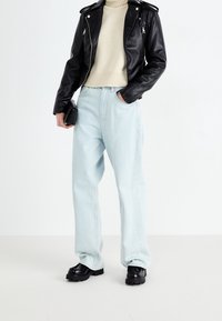 Schwarze Lederbikerjacke, cremefarbener Strickpullover, hellblaue weit geschnittene Jeans und schwarze klobige Schuhe. Hält eine kleine schwarze Tasche.