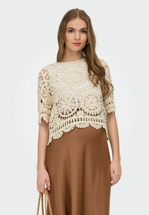 Tânără cu părul lung, purtând un top bej croșetat cu mâneci scurte peste un strat alb și o fustă maro, ținând o geantă țesută.