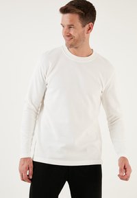 Weißes Langarmhemd mit gerippter Textur und runder Ausschnittform, mit lockerer Passform und geradem Saum. Getragen mit dunklen Hosen.