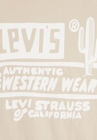 Camiseta beige con gran texto impreso en blanco que dice "Levi's", "Ropa Western Auténtica" y "Levi Strauss & Co." con un gráfico de cacto.