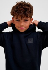 Svart hoodie med framficka, texturerat tyg och en liten, upphöjd logotypmärke på bröstet. Huvan är uppfälld och ramar in ansiktet.