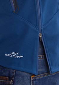 Gros plan d'une veste bleue GORE WINDSTOPPER avec fermeture éclair avant partiellement ouverte, portée sur un jean bleu.