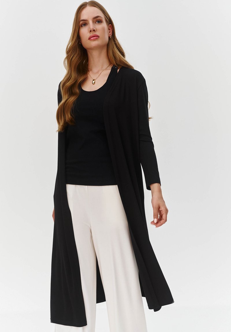 Długi czarny kardigan wykonany z miękkiego materiału, z otwartym przodem, długimi rękawami i luźnym krojem, noszony na czarnym topie i białych spodniach.