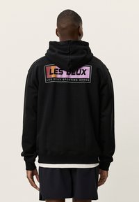 Μαύρο φούτερ με κουκούλα που έχει ένα μεγάλο πολυχρωματικό λογότυπο στην πλάτη με το κείμενο «LES DEUX» και «LES DEUX SPORTING GOODS» σε λευκό.