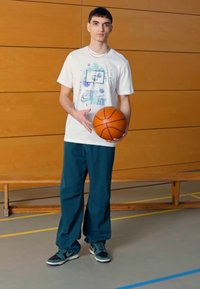 Camiseta blanca con un gráfico de baloncesto, letras en azul verdoso y acentos en morado; pantalones en azul profundo; sosteniendo un balón de baloncesto; zapatillas negras y azul verdoso.