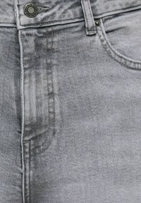 Gros plan sur un jean en denim gris délavé montrant les coutures, la poche avant, la passants de ceinture et un bouton en métal marqué "Blue Denim 1965".