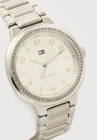 Ασημένιο ρολόι χειρός Tommy Hilfiger με στεφάνι επενδυμένο με κρύσταλλα, λευκό καντράν, ασημένιους δείκτες ώρας και τρεις δείκτες που δείχνουν 12:13:59.