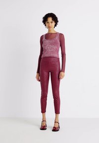 Débardeur rose ajusté superposé sur une chemise à manches longues transparente, associé à des leggings bordeaux brillants avec un motif subtil, portés avec des chaussures rouges.