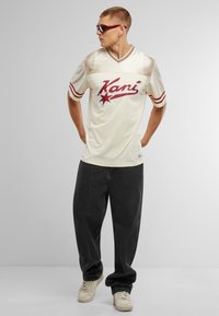 Kreminė tinklinė marškinė su raudonais akcentais ir "Kani" logotipu, derinami su tamsiai pilkomis plačiomis kelnėmis ir baltais sportbačiais. Modelis dėvi akinius nuo saulės.