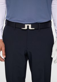 Zwarte riem met een zilveren metalen JL-buckle, gedragen over marineblauwe broeken. Het model draagt een lichtblauw shirt en een witte golfschoen aan de linkerhand.