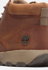 Brązowe skórzane botki za kostkę o teksturowanym wykończeniu, z metalowym logo w kształcie drzewa i beżową gumową podeszwą, z detalicznym przeszyciem wzdłuż szwów.