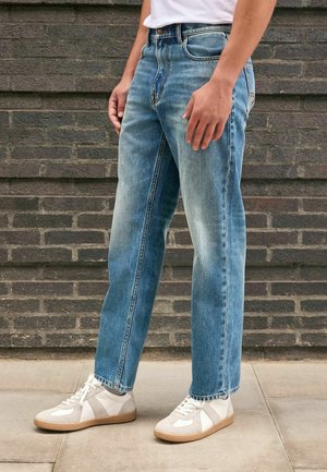 Homme portant un jean droit bleu clair et des baskets blanches, debout sur un trottoir avec un mur en briques sombre en arrière-plan.