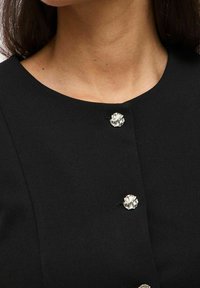 Svart plagg med rund halsringning och tre dekorativa silverfärgade knappar med en texturerad, blommiliknande design. Mjuk tygtextur.