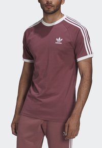 Bordo sporta t-krekls ar baltu apkakli un piedurkņu apmalēm, kurā ir trīs baltas svītras uz pleciem un neliels logotips uz krūtīm.