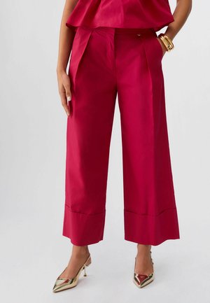 Donna che indossa pantaloni rossi a vita alta e gamba larga con risvolti, tacchi dorati a punta e braccialetti d'oro a un polso.