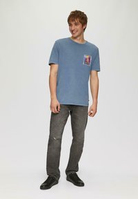 Blauwe katoenen T-shirt met een borstzak met een kleurrafisch motief. Draag het met versleten zwarte spijkerbroeken en zwarte sneakers.